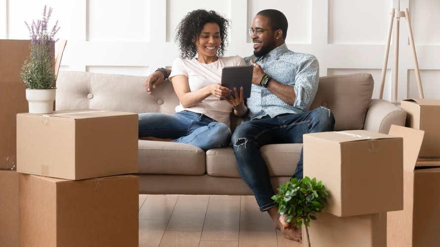 Pareja de hombres y mujeres sentados en un sofá mirando una tableta mientras está rodeado de cajas móviles soluciones de préstamos hipotecarios