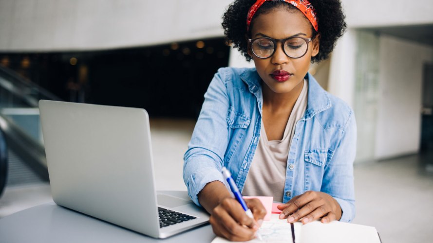 Mujer negra toma notas en papel mientras está sentada frente a una computadora portátil: emprendedora de la pandemia de pequeñas empresas